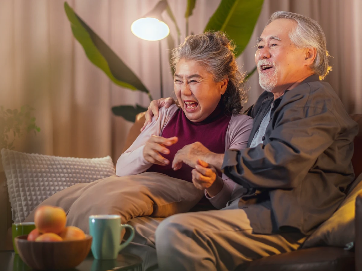 Elderly Asian couple with excited expressions watching engaging brand content on television.